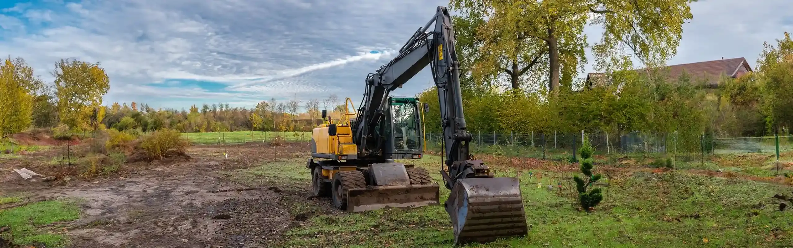 Wheeled excavator parked on cleared ground beside a garden boundary.